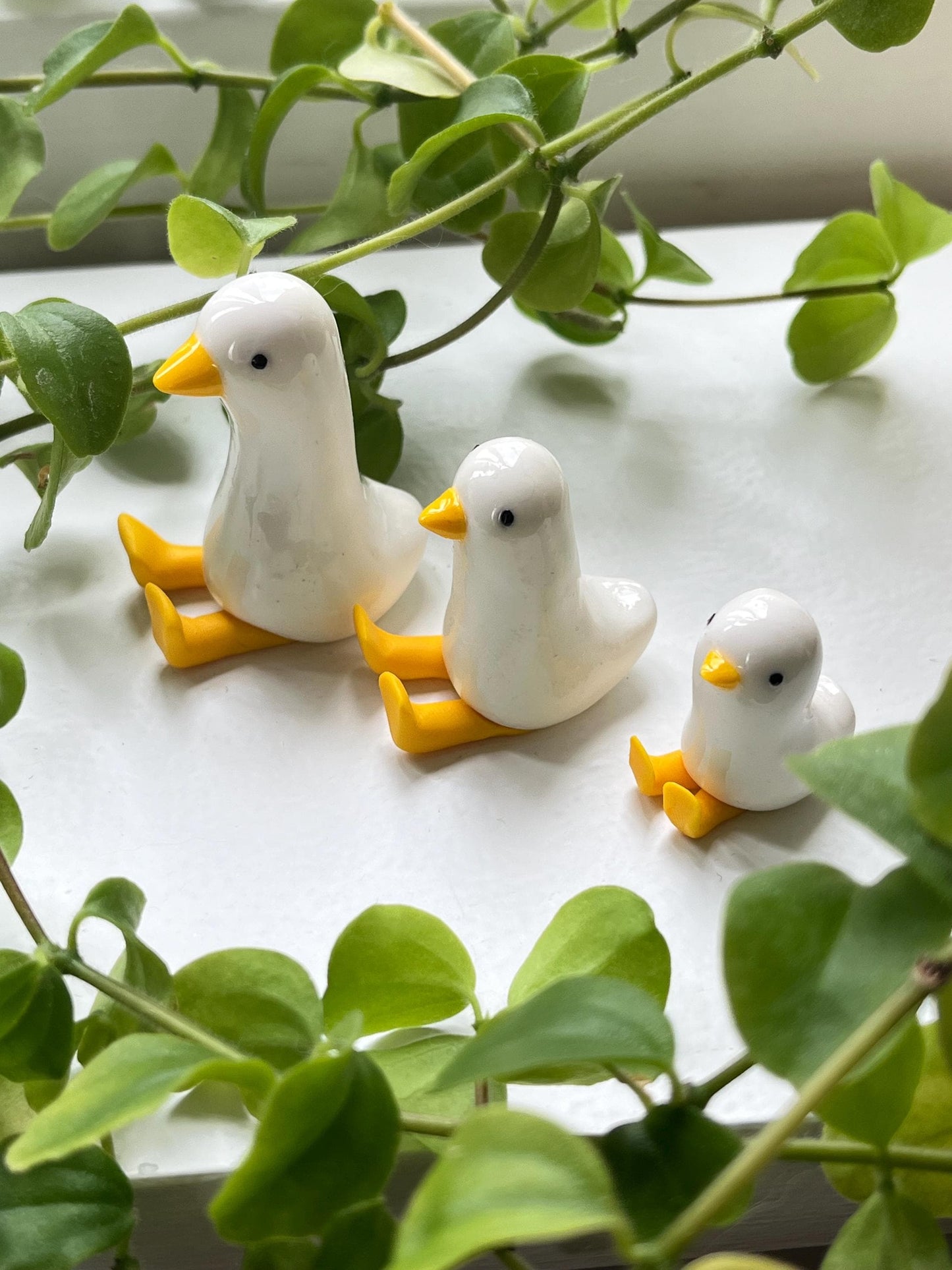 Three white duck figurines with yellow feet on a white surface surrounded by green leaves.